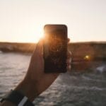 person holding black phone beside body of water under white sky during golden hour