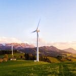 A wind turbine stands tall against a mountainous backdrop under a vibrant sky at sunset.